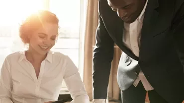 Two colleagues working together at a desk with sunlight streaming through a window.
