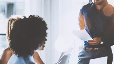 People in an office meeting, discussing documents in bright sunlight.