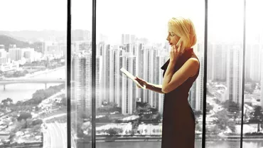 Person standing by large window, holding a tablet with a cityscape in the background.