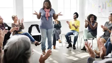 Person standing in a room surrounded by people clapping and smiling.