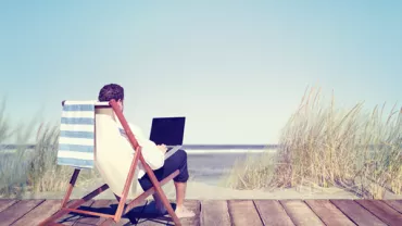 Person relaxing on a deck chair by the beach with a laptop.