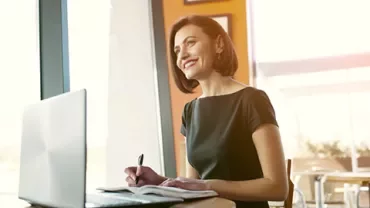 Person smiling while working on a laptop near a window.