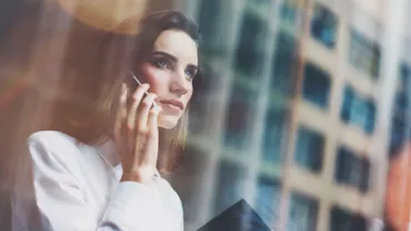 Person looking thoughtful while talking on a phone, standing by a window.