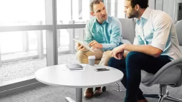 Two people in an office discuss something on a tablet at a round table.