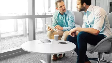 Two people in an office discuss something on a tablet at a round table.