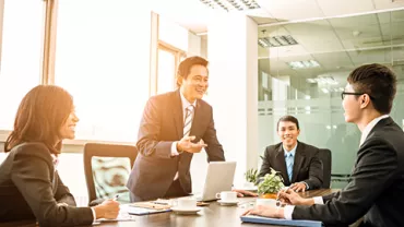 Group of people in a meeting, with one person standing and talking, while others listen and take notes.