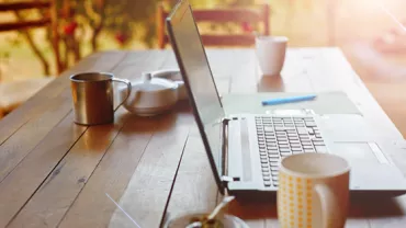 Open laptop on a wooden table with mugs and a teapot in a sunlit setting.