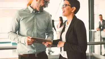 Two colleagues laughing while discussing work with a tablet in an office setting.