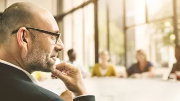 A person wearing glasses in a meeting room, surrounded by colleagues at a large table.