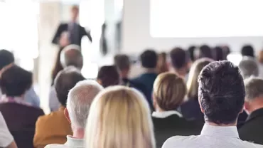 A large audience listens to a presentation in a bright conference room.