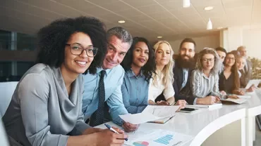 A diverse group of people smiling and holding papers at an office meeting.