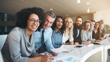 A diverse group of people smiling and holding papers at an office meeting.
