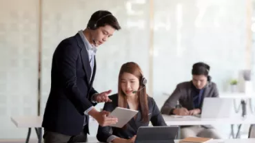 Two people in an office setting, one standing with a tablet, the other seated at a desk with a laptop.