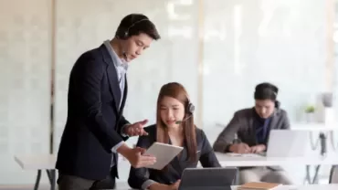 Two people in an office setting, one standing with a tablet, the other seated at a desk with a laptop.