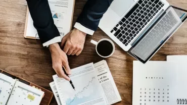 Person reviewing financial charts at a wooden desk with laptop and coffee.