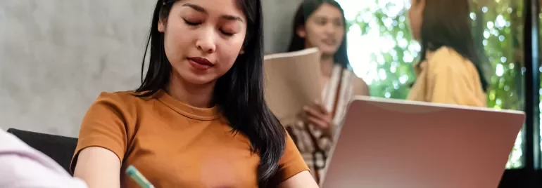 Person focuses on writing at a desk, with two others talking in the background.