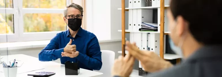 Two people wearing masks, communicating in sign language in an office.