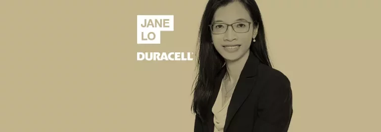 Person in a formal outfit with glasses, smiling against a solid background.