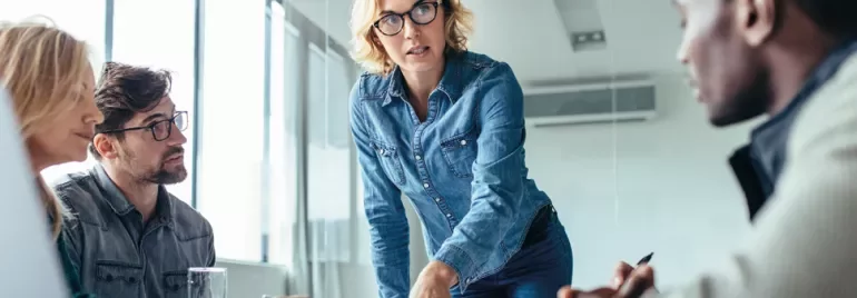 Person in denim speaking at a meeting, with others listening attentively around a table.