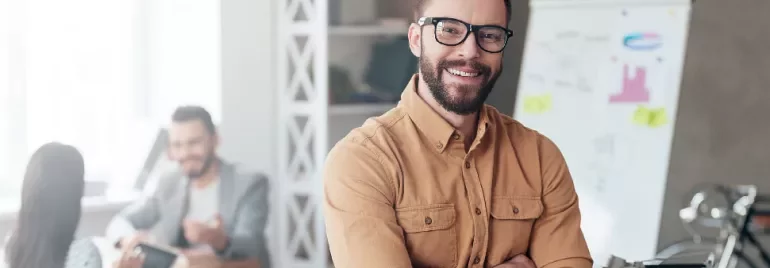 Person smiling, standing in an office with a whiteboard and two people in the background.