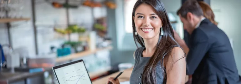 Person smiling at camera while holding a pen, with a laptop displaying graphs in an office setting.