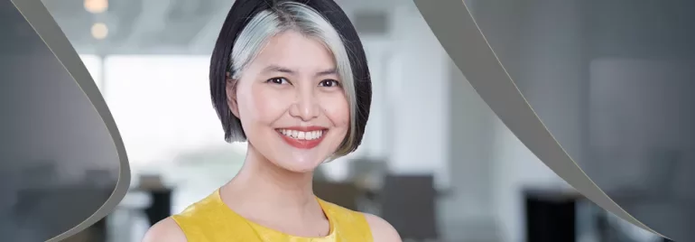 Person smiling with short hair, wearing a yellow top, in a blurred office setting.