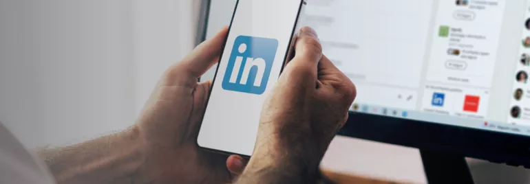 Hands holding a smartphone displaying the LinkedIn logo, with a computer screen in the background.