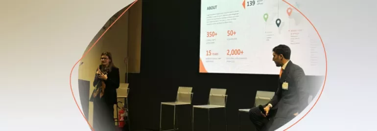 Two people participating in a presentation, with one speaking on stage and a large slide displayed behind them.