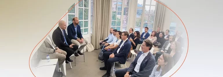 Group attending a discussion in a bright room, with several people seated in rows listening to a small panel at the front.