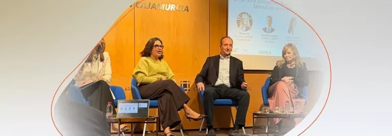Panel of speakers seated on stage during an event, with a presentation screen and wooden backdrop behind them.