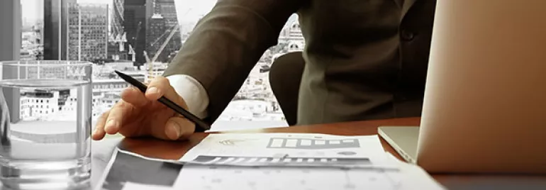 Person in a suit working at a desk with a laptop and papers, cityscape visible through the window.