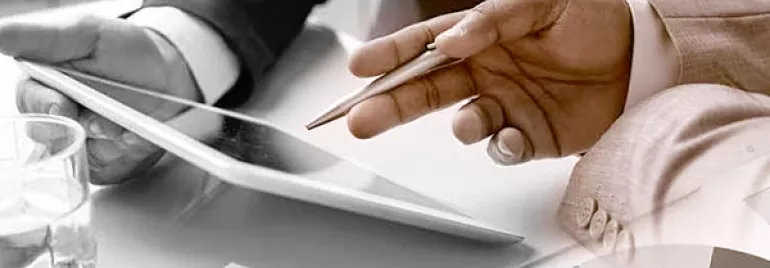 Hands holding a tablet and pen above paperwork with pie charts and graphs.