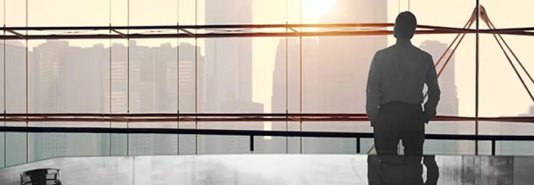 Person looking at city skyline through large office window, desk in foreground.