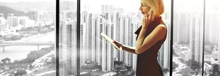 Person standing by large window, holding a tablet with a cityscape in the background.