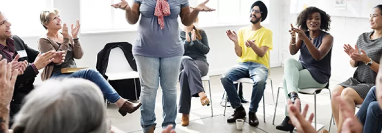 Person standing in a room surrounded by people clapping and smiling.