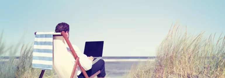 Person relaxing on a deck chair by the beach with a laptop.