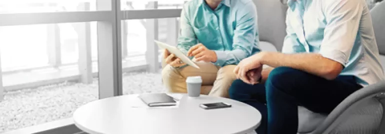 Two people in an office discuss something on a tablet at a round table.