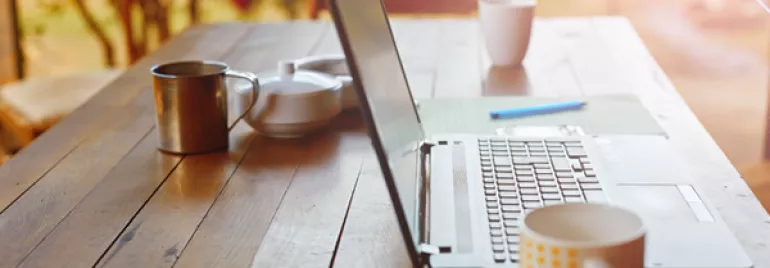 Open laptop on a wooden table with mugs and a teapot in a sunlit setting.
