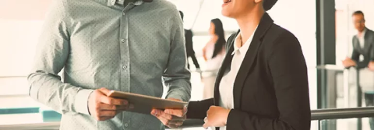 Two colleagues laughing while discussing work with a tablet in an office setting.