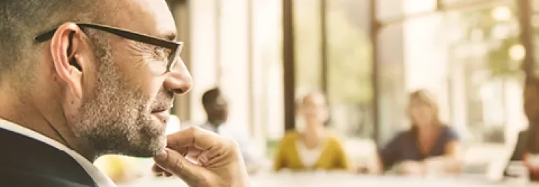 A person wearing glasses in a meeting room, surrounded by colleagues at a large table.