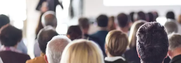 A large audience listens to a presentation in a bright conference room.