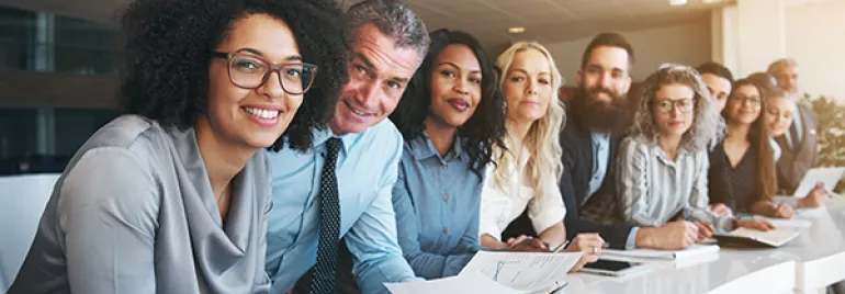 A diverse group of people smiling and holding papers at an office meeting.