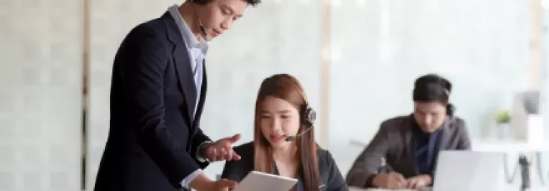 Two people in an office setting, one standing with a tablet, the other seated at a desk with a laptop.