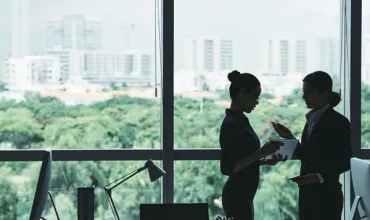 Two colleagues discussing work by an office window with a city view.