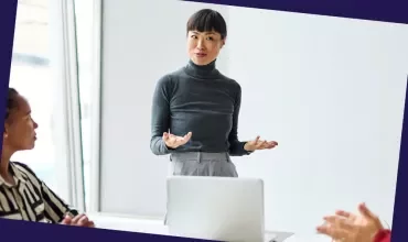 Person presenting to colleagues during a meeting, standing beside a laptop in a bright office.