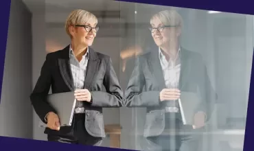 Person in business attire holding a laptop while standing beside a glass wall in an office.