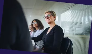 Person wearing glasses seated in a meeting room, with colleagues and office blinds in the background.