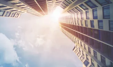 Upward view of tall buildings with sunlight peeking through a partly cloudy sky.