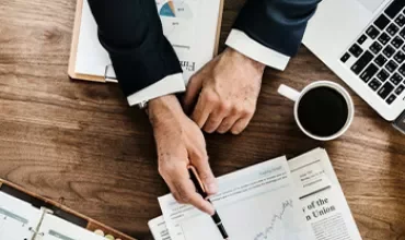 Person analyzing financial charts with a pen at a wooden desk.