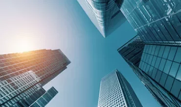 Looking up at modern skyscrapers against a clear blue sky with sunlight peeking through.